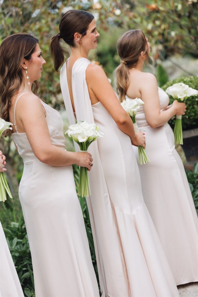 Three women in light-colored dresses stand in a row outdoors, each holding a bouquet of white flowers.