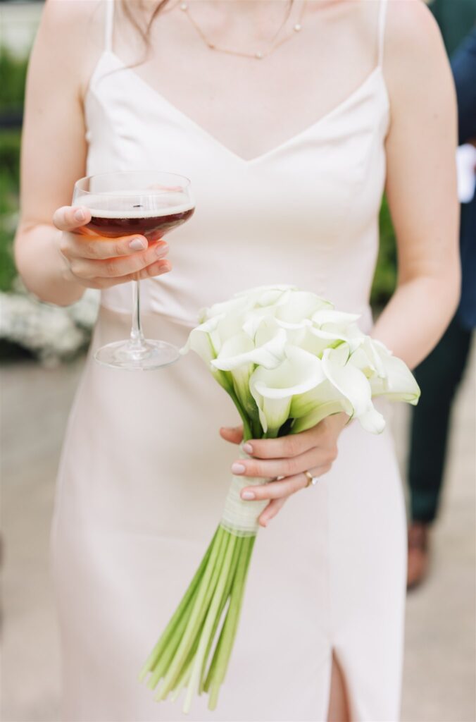 A woman in a white dress holds a bouquet of white calla lilies in one hand and a glass of red wine in the other.