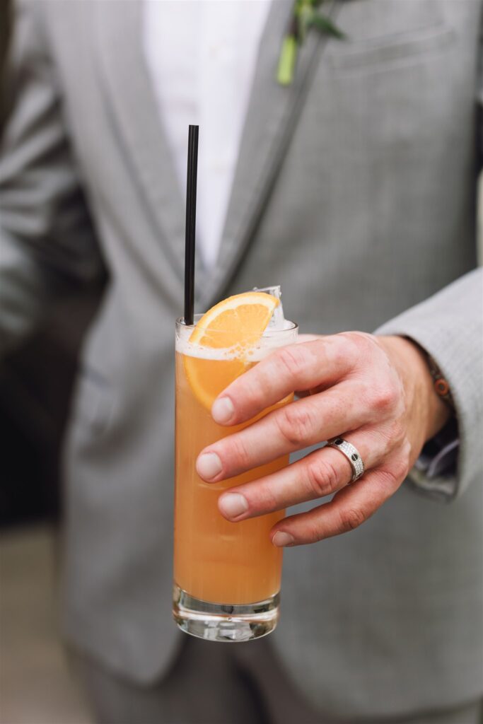 Person in a light gray suit holding a tall glass of orange-colored drink with ice, a slice of orange, and a black straw.