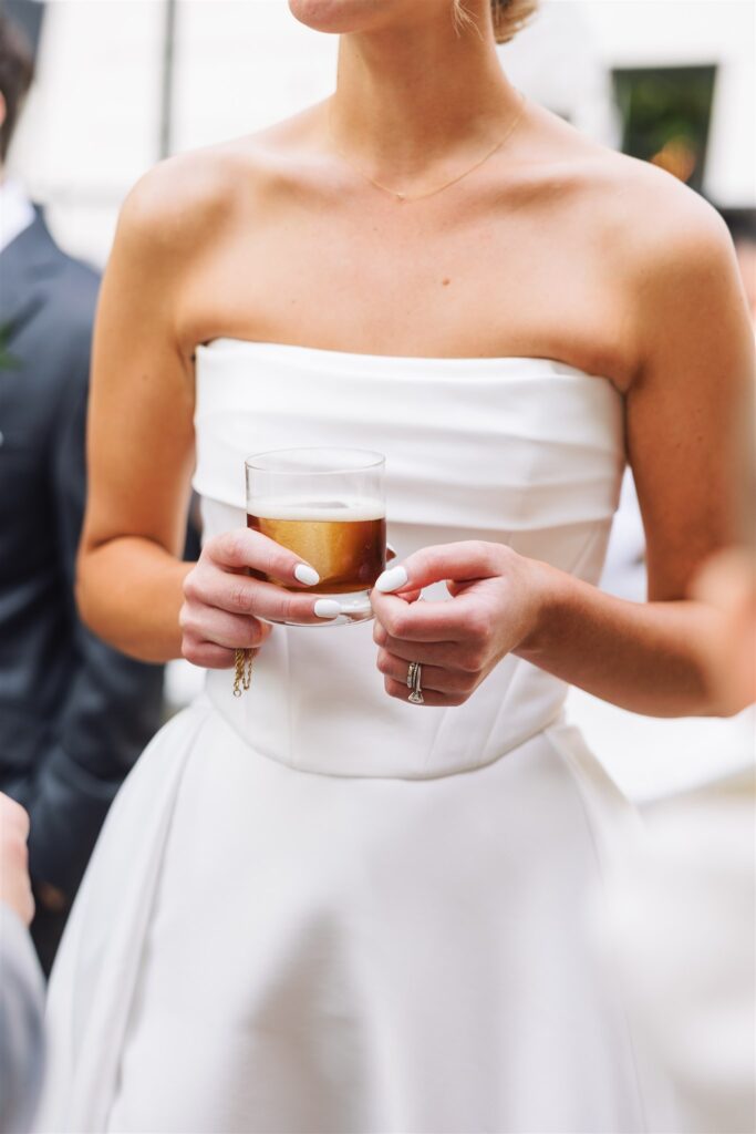 A woman in a strapless white dress holds a cocktail glass with a foamy drink.