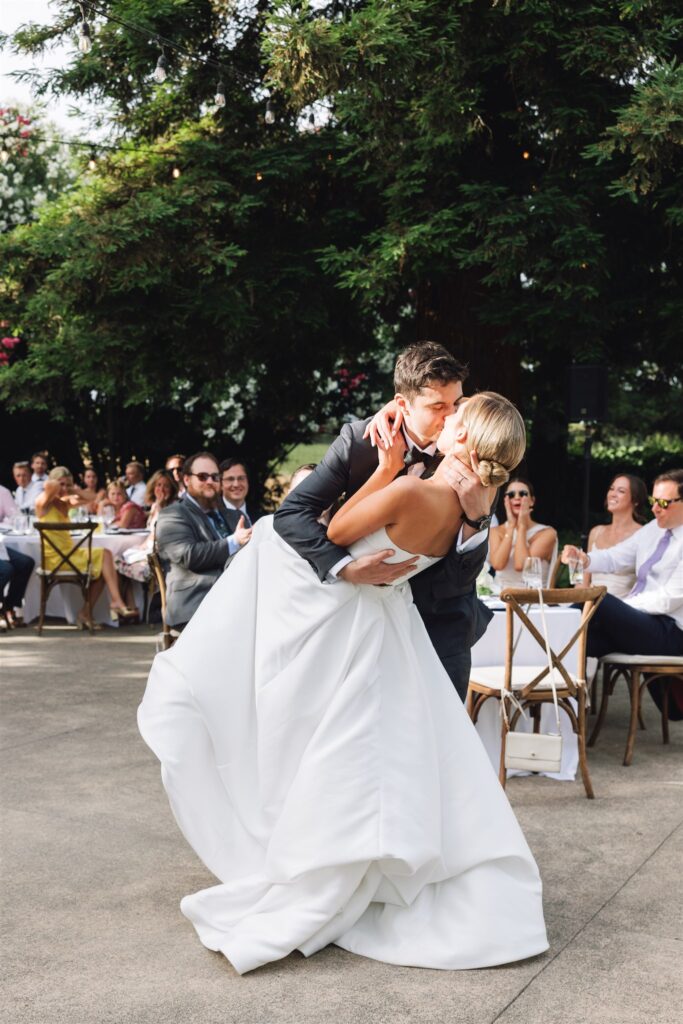 A groom in a black suit dips and kisses a bride in a white dress on an outdoor dance floor, with seated guests watching in the background.