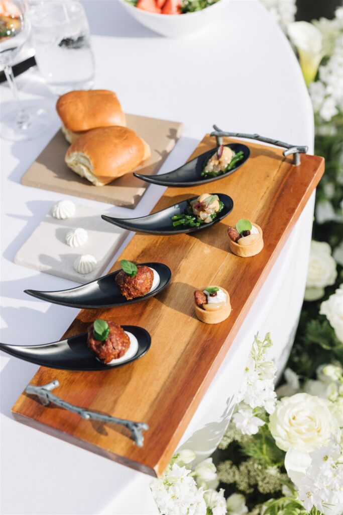A wooden serving board with assorted bite-sized appetizers on black spoons and pastry rounds, next to bread rolls on a white table with floral decor.