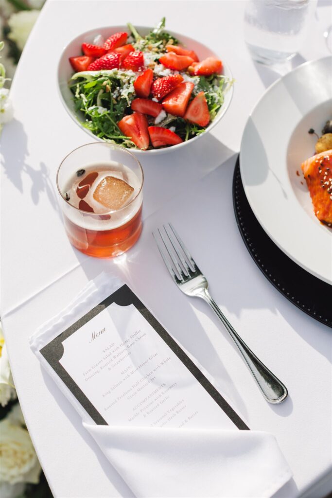 A table setting with a salad topped with strawberries, a drink with ice, a fork, a menu, and a plate of salmon on a white tablecloth.