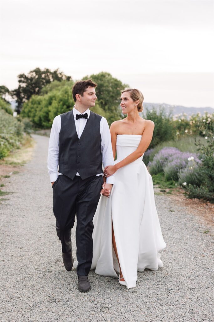 A couple in formal wedding attire walks hand in hand on a gravel path surrounded by greenery, looking at each other.