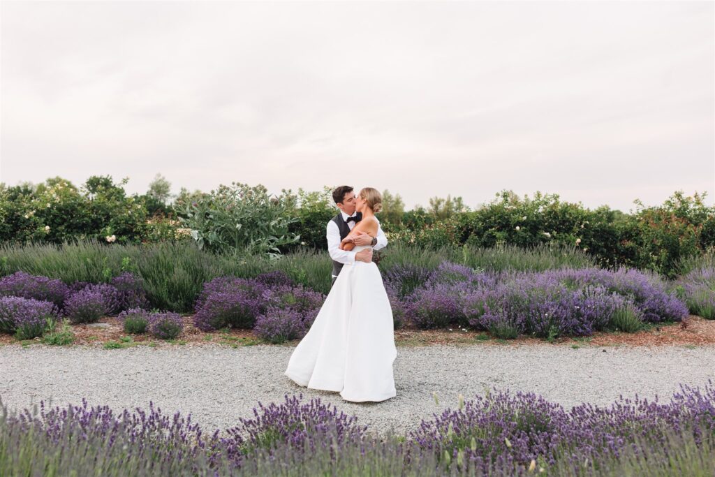 A couple in wedding attire embraces on a gravel path surrounded by blooming lavender and greenery under a cloudy sky.