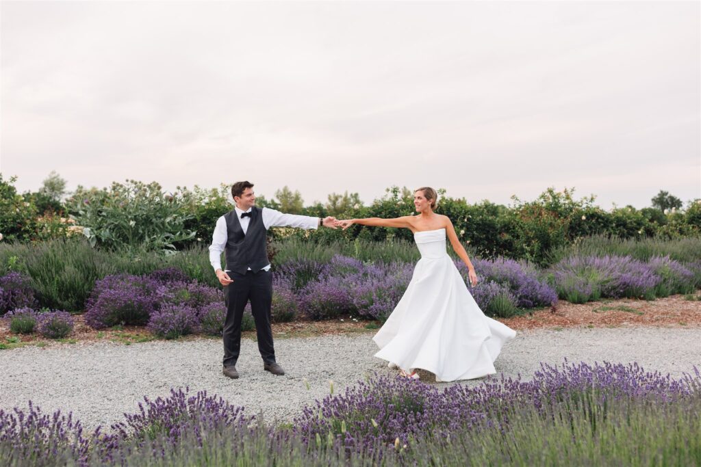A bride and groom hold hands and dance on a gravel path surrounded by lavender bushes and greenery under a cloudy sky.