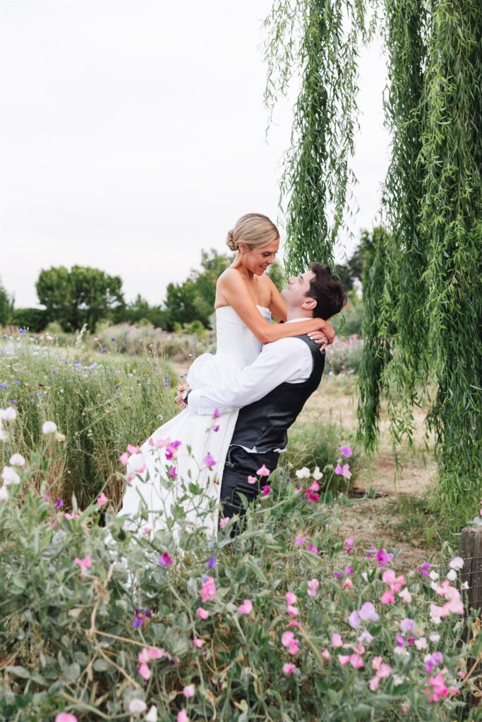A groom lifts a bride in a white dress among wildflowers and greenery, both smiling under a weeping willow tree in an outdoor setting.