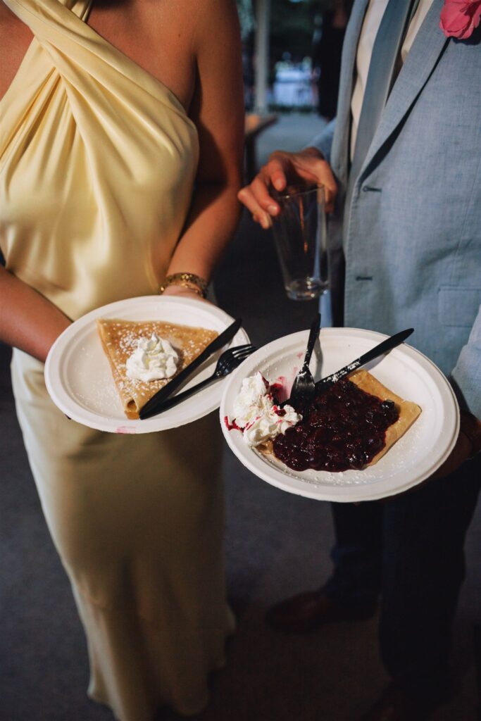 Two people holding plates with crepes; one topped with whipped cream, the other with whipped cream and berry sauce. One person holds an empty glass. Both are dressed formally.