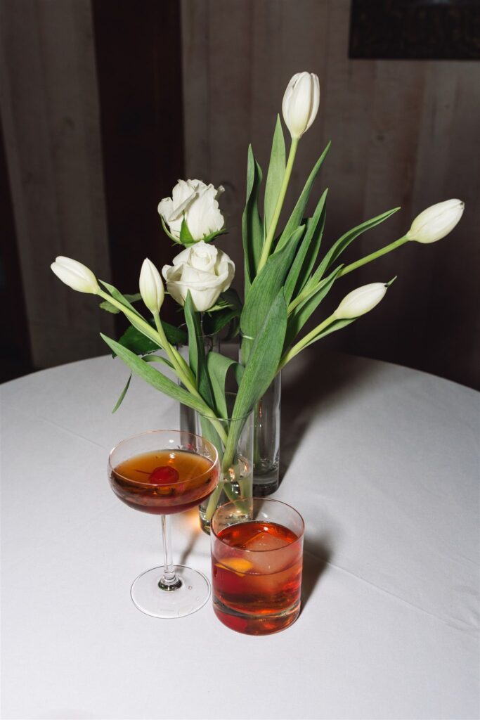 A round table with a white tablecloth holds a vase of white tulips and roses, a coupe glass with a dark cocktail and cherry, and a rocks glass with an orange-red drink and ice.