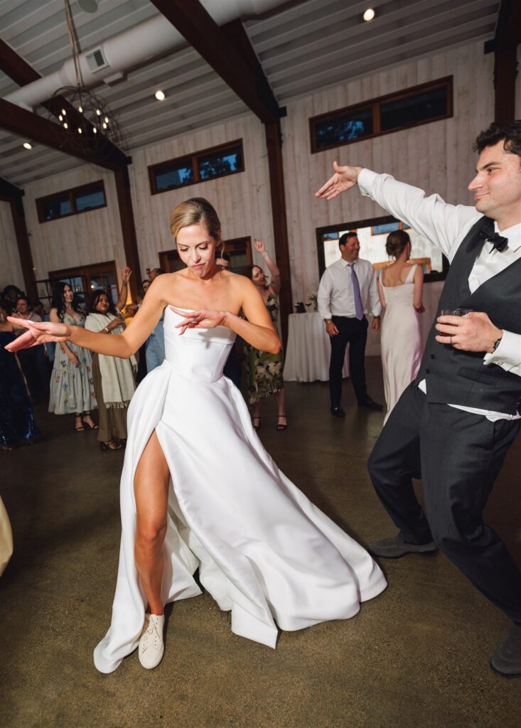 A bride in a white dress and sneakers dances energetically with guests at an indoor wedding reception.