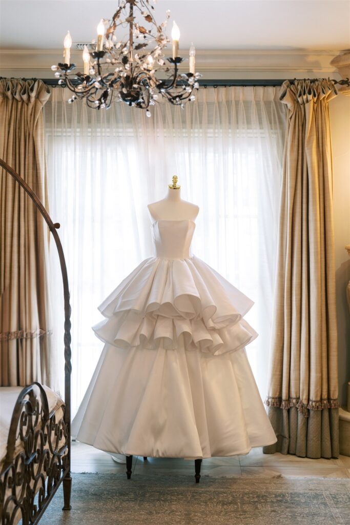 A white strapless ball gown with a tiered ruffled skirt displayed on a dress form in a room with large windows, beige curtains, and a chandelier overhead.