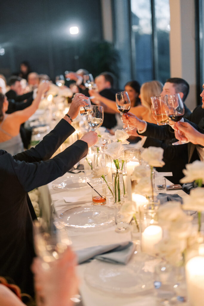 A group of people seated at a long table raise their glasses in a toast, surrounded by candles, flowers, and soft lighting.