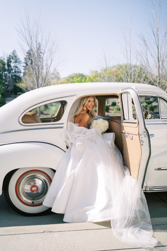A bride in a white gown and veil steps out of a vintage white car holding a bouquet on a sunny day.