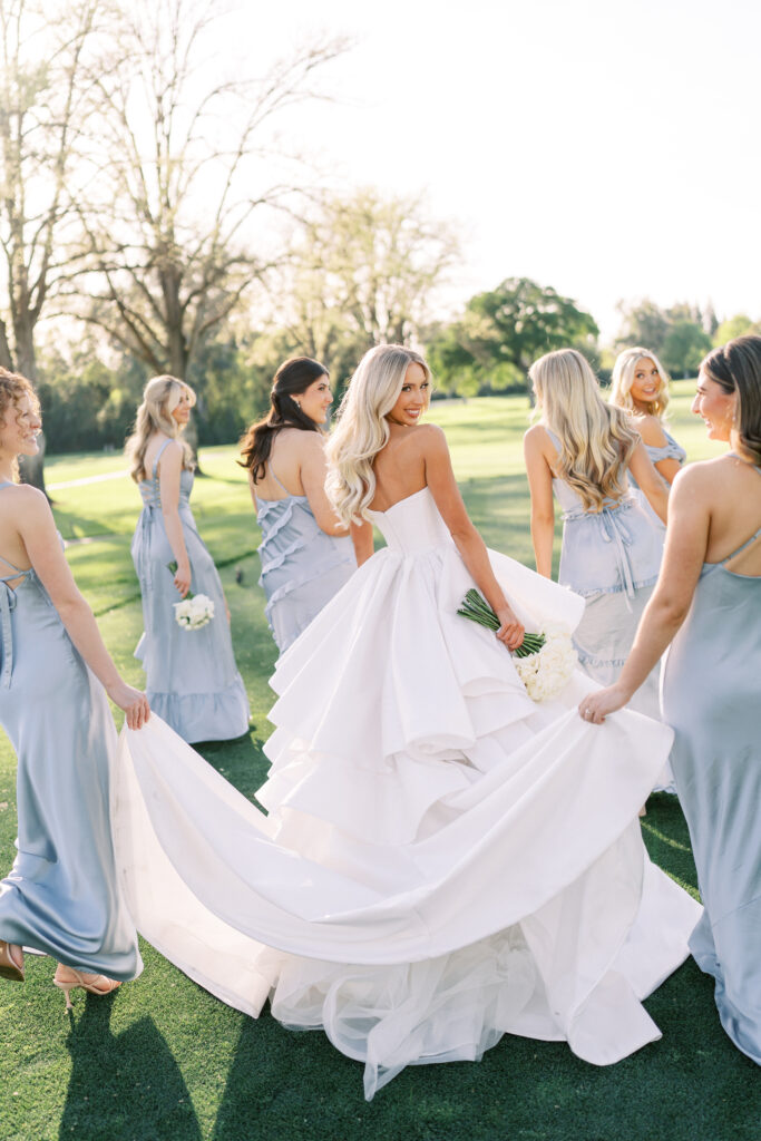 A bride in a white gown holds a bouquet and looks back while walking on grass, surrounded by bridesmaids in light blue dresses.