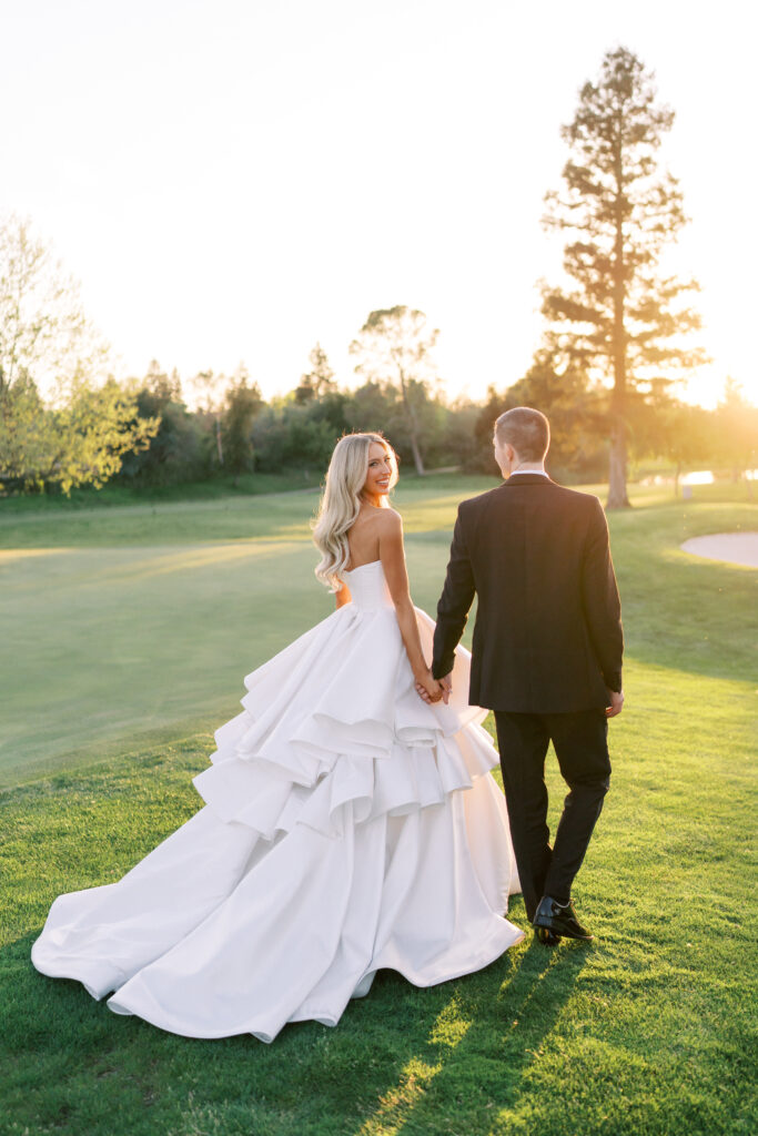 A bride in a white gown and a groom in a black suit hold hands and walk on a grassy field at sunset.
