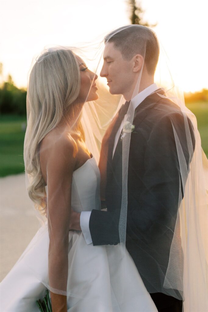 A bride and groom stand close together outdoors at sunset, wrapped in a veil, looking into each other's eyes.