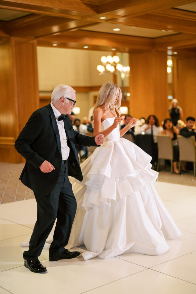 An older man in a black tuxedo and a woman in a white wedding gown dance together on a white floor at an indoor event with seated guests in the background.