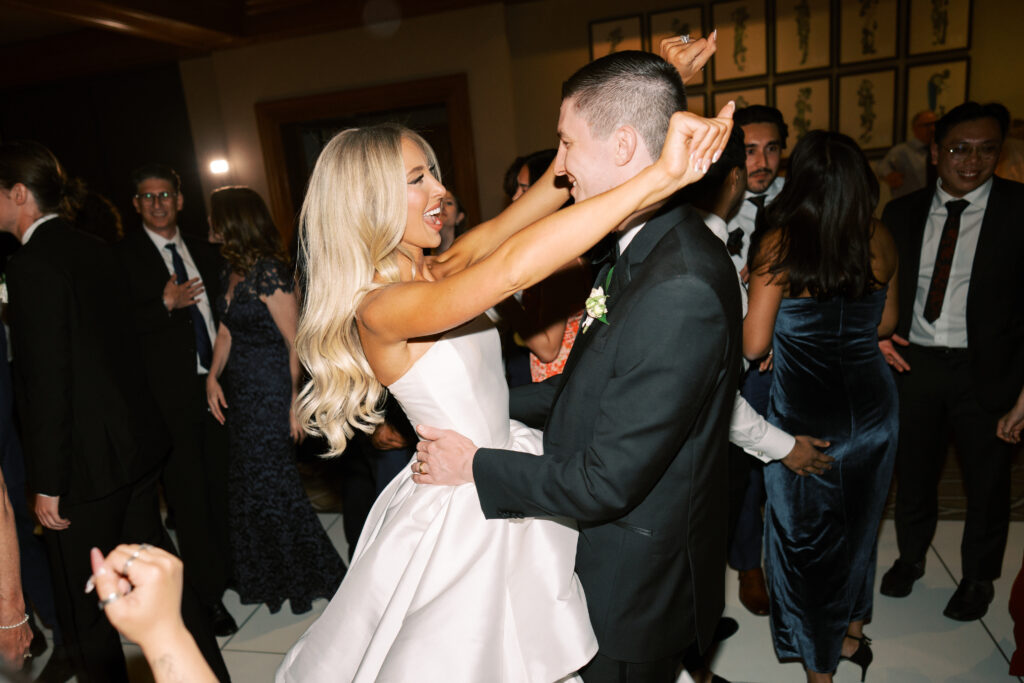 A bride and groom dance together at their wedding reception, planned by a Northern California wedding planner, surrounded by guests in formal attire—many joining them on the dance floor.