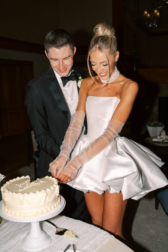 A bride and groom, both smiling, cut a white frosted cake together at their wedding reception.