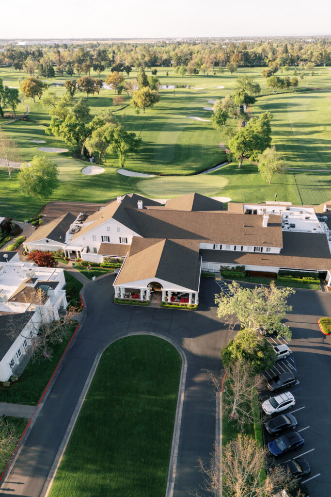 Aerial view of a clubhouse building with parked cars in front, surrounded by landscaped golf course and green fairways.