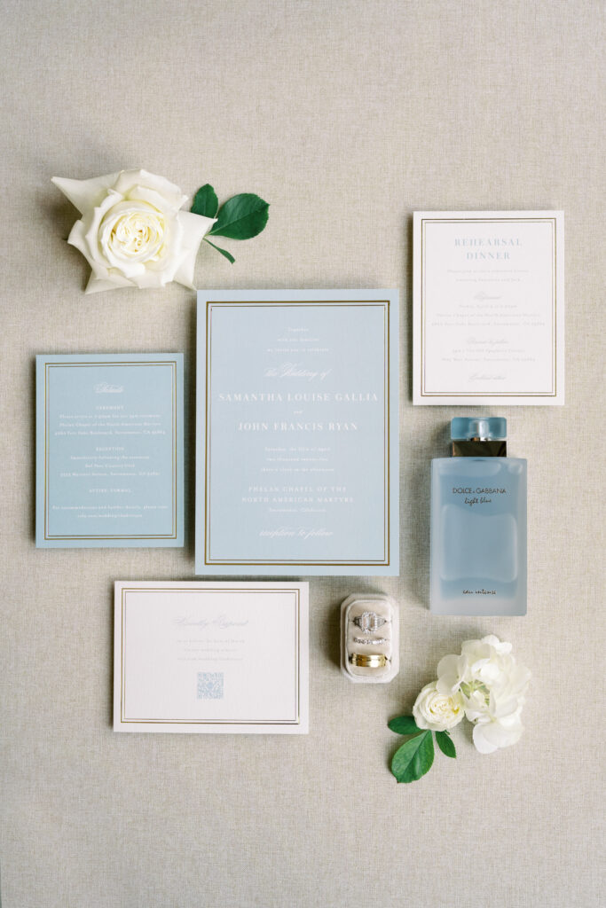 A flat lay of wedding stationery, including invitations, RSVP cards, a rehearsal dinner card, perfume, two white roses, and a small box with rings, arranged on a neutral fabric background.
