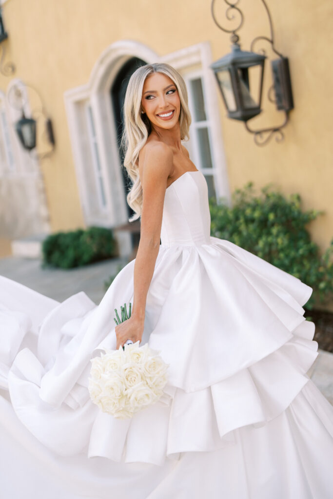 A bride in a strapless white wedding dress holds a bouquet of white roses and smiles while standing outdoors in front of a yellow building.