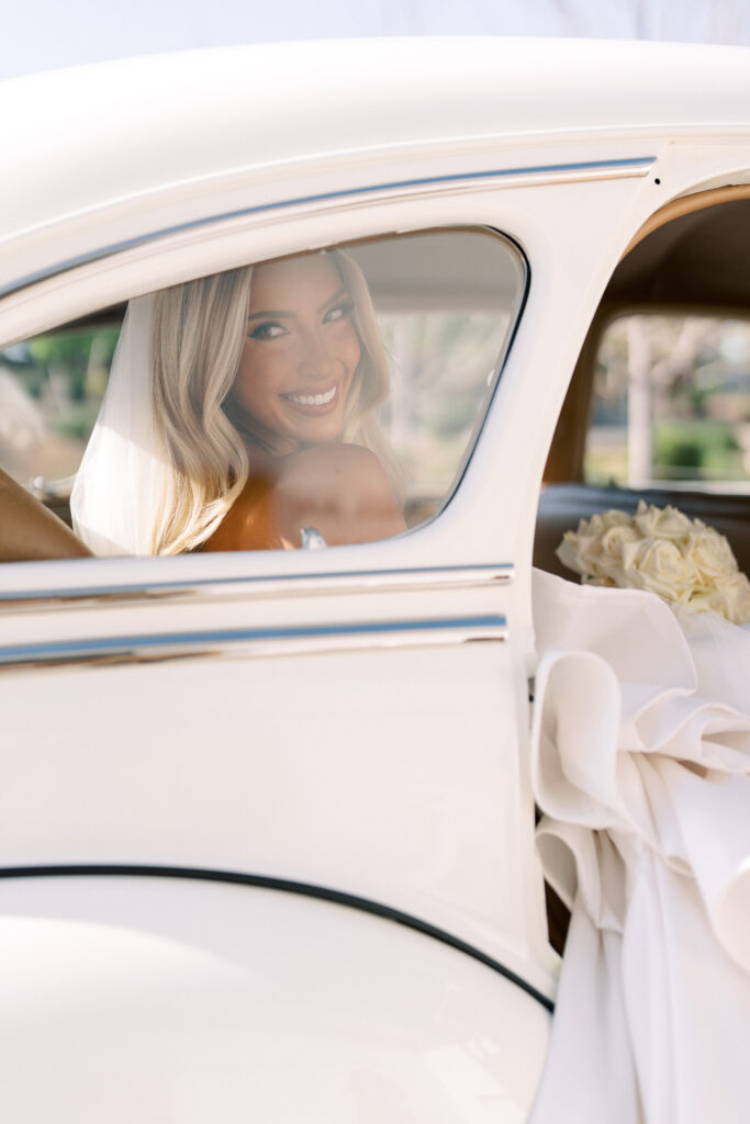 Bride with long blond hair smiles while sitting in the backseat of a classic white car, holding a bouquet of white flowers and wearing a veil.