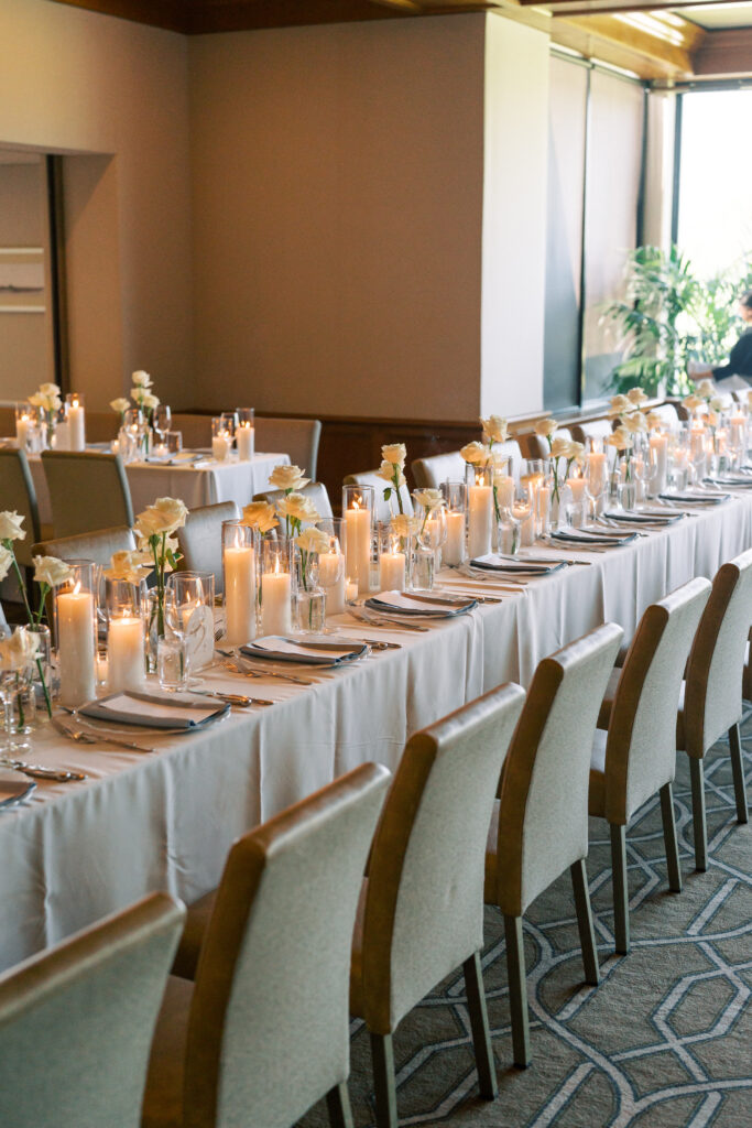 A long dining table set with white linens, glassware, candles, and white flowers, surrounded by beige chairs in a well-lit room.
