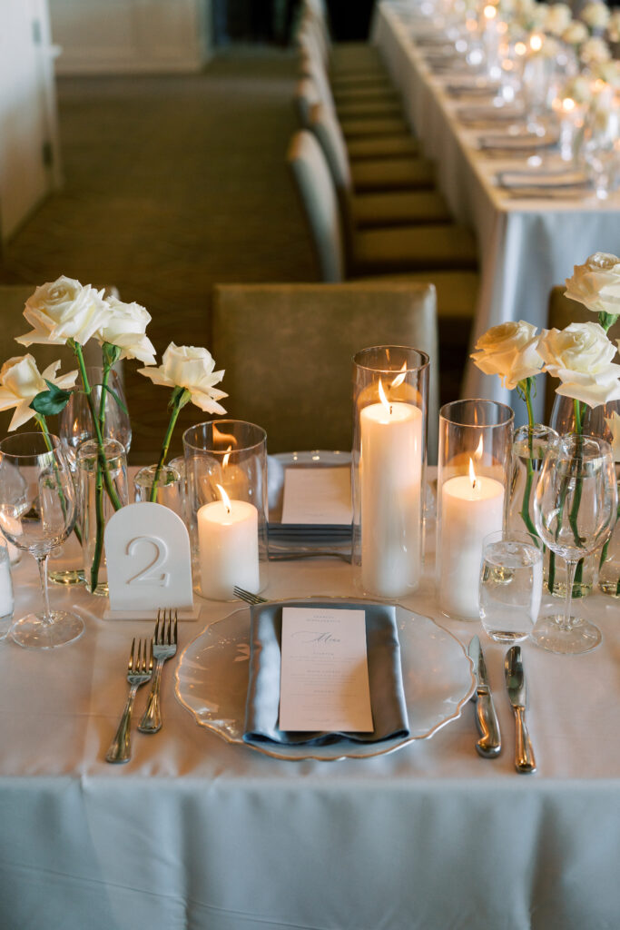 Elegant table setting with white roses, candles, glassware, silver cutlery, a translucent charger plate, and a table number "2" on a white tablecloth.