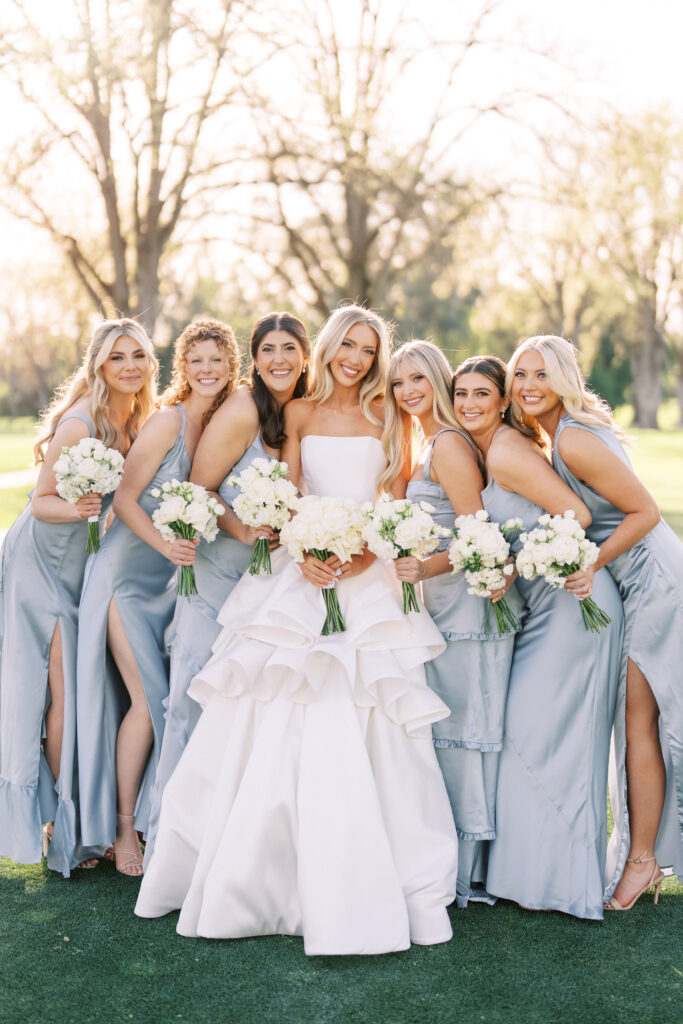 A bride in a white gown stands outdoors with six bridesmaids in light blue dresses, all holding bouquets and smiling at the camera.