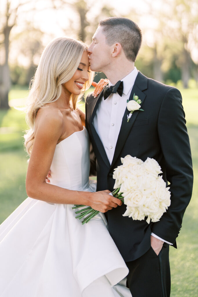A bride in a strapless white gown holds a bouquet of white roses as a groom in a black tuxedo kisses her forehead outdoors.