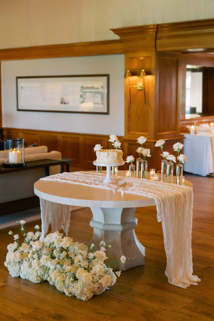 A round table with a white cloth, white floral arrangements, a small cake, and vases with white flowers, set in a wood-paneled room.