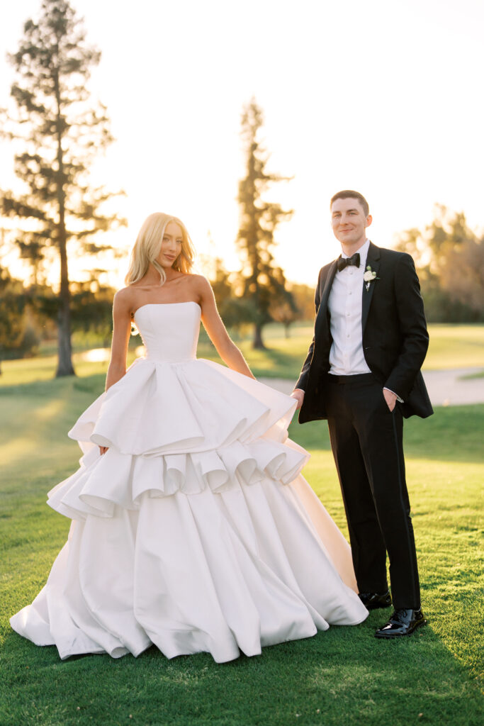 A bride in a strapless white gown with layered ruffles stands beside a groom in a black tuxedo on a grassy lawn at sunset.