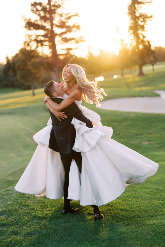 A groom lifts and spins the bride on a golf course at sunset, both smiling and dressed in formal wedding attire.