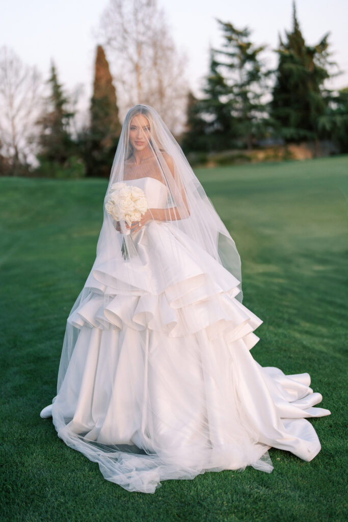 A bride in a strapless white wedding gown with a layered skirt and a long veil holds a bouquet of white flowers while standing on a grassy area outdoors.