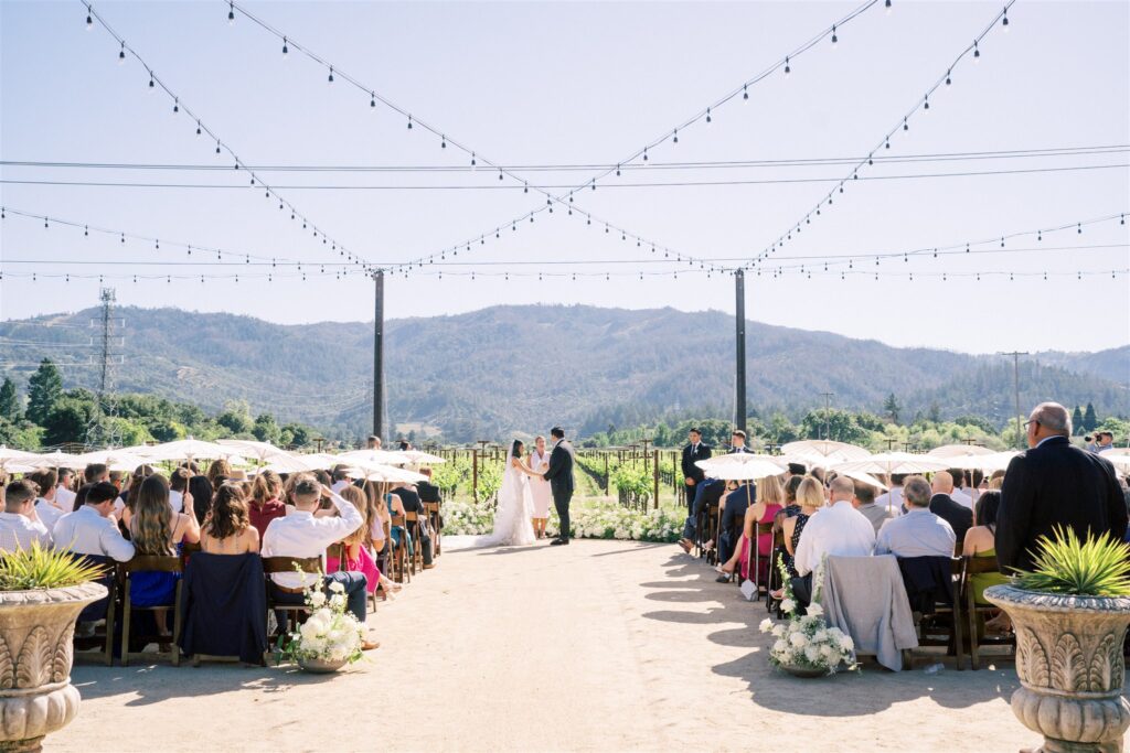 An outdoor wedding ceremony with guests seated on both sides, a couple standing at the altar, string lights overhead, and mountains visible in the background.