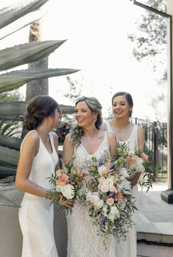 Three women in white dresses stand together outdoors, smiling and holding bouquets of flowers. Succulents and a fence are visible in the background.
