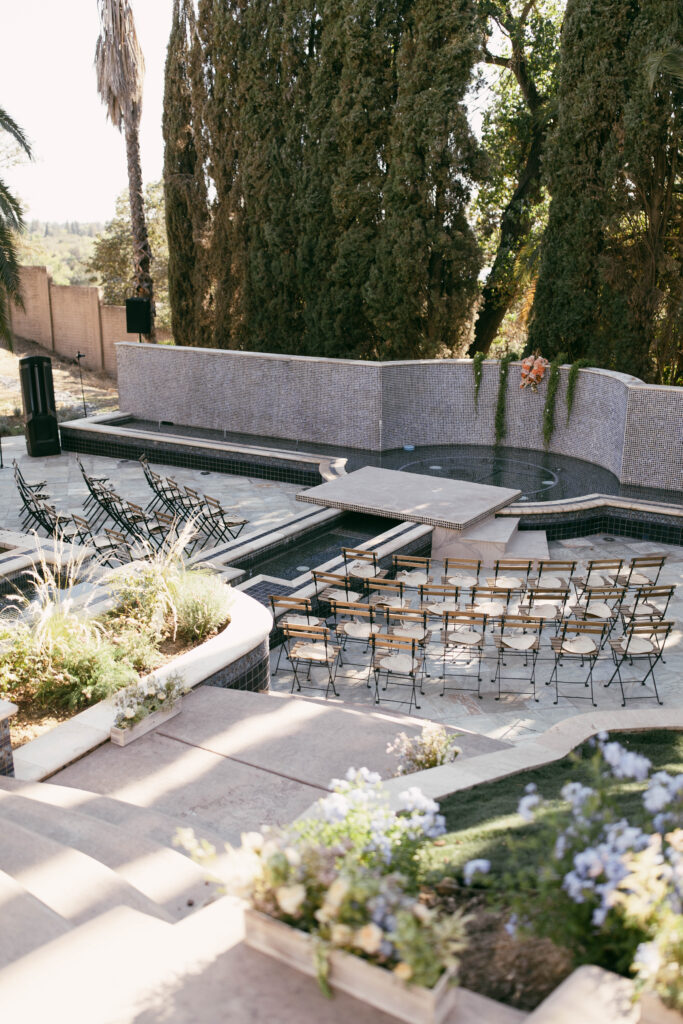 Outdoor wedding ceremony setup with rows of folding chairs facing a small stage, surrounded by tall trees and stone walls, with floral decorations in the foreground.