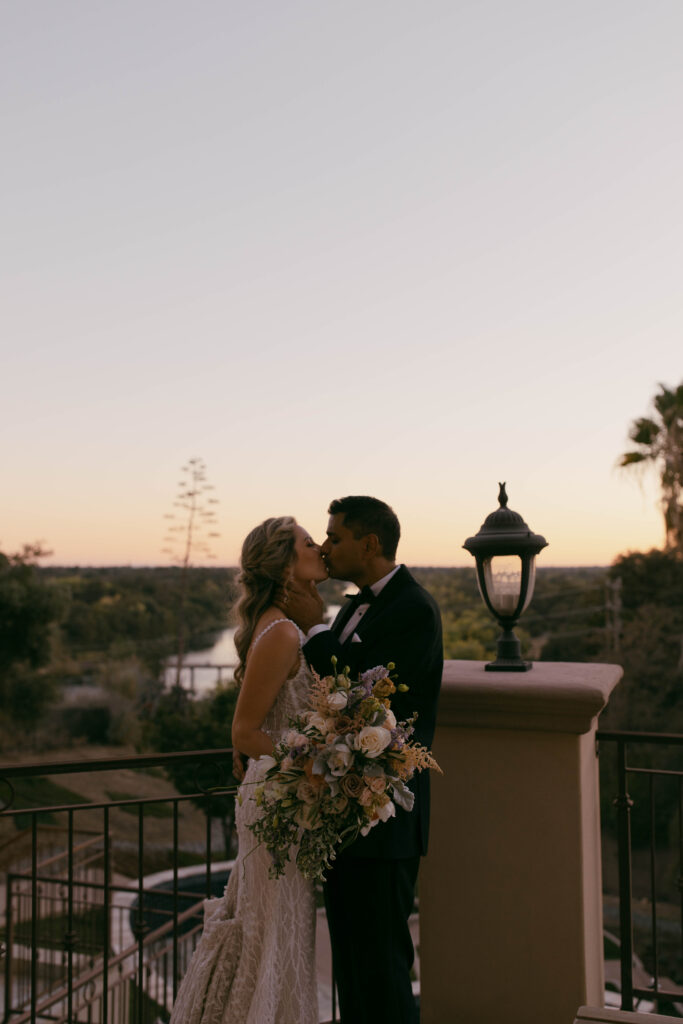 A bride and groom stand close together outdoors at sunset, the bride holding a bouquet of flowers, with trees and a lamp post in the background.
