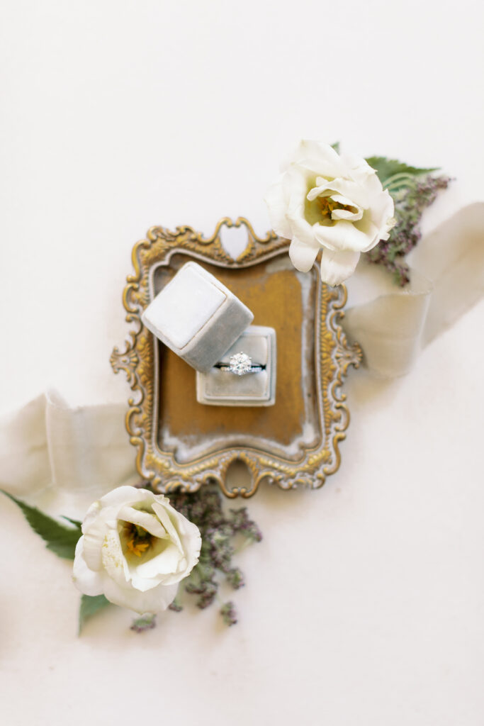 A diamond ring in a gray velvet box rests on a decorative gold tray, surrounded by white flowers and a beige ribbon on a white background.