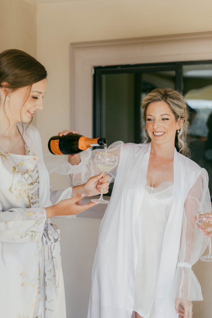 Two women in light-colored robes, one pouring champagne into a glass held by the other, both smiling indoors near a window.