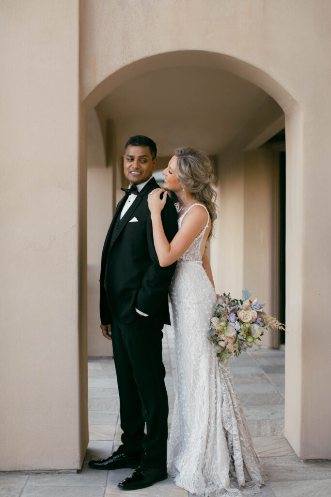 A bride in a white lace dress stands behind a groom in a black tuxedo under an archway, holding a bouquet and smiling.