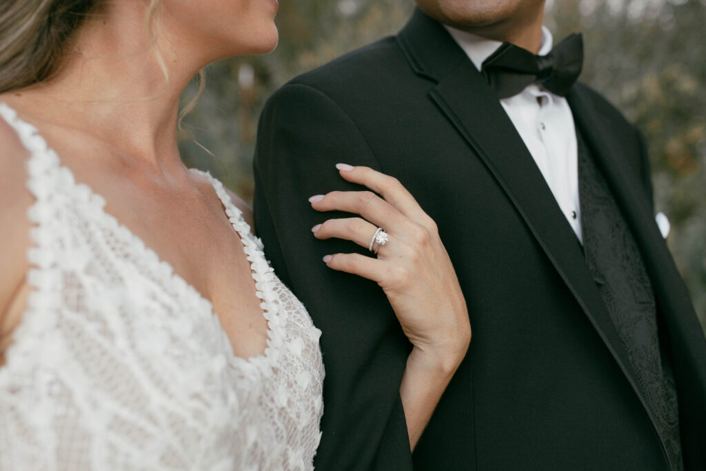 Close-up of a bride holding the arm of a groom in a black tuxedo, showing her lace dress and diamond ring.