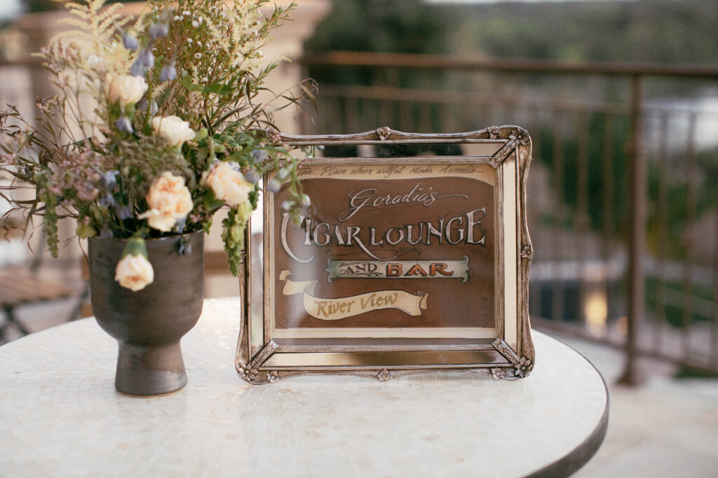 A framed sign reading "Gerald's Cigar Lounge and Bar River View" sits on a round table next to a vase of flowers, with a railing and natural landscape in the background.
