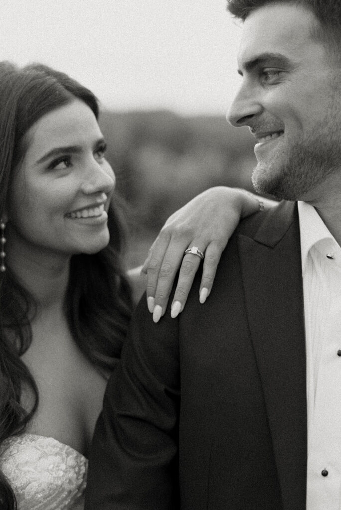 A woman in a strapless dress smiles at a man in a suit, resting her hand with a ring on his shoulder. Both are looking at each other closely.