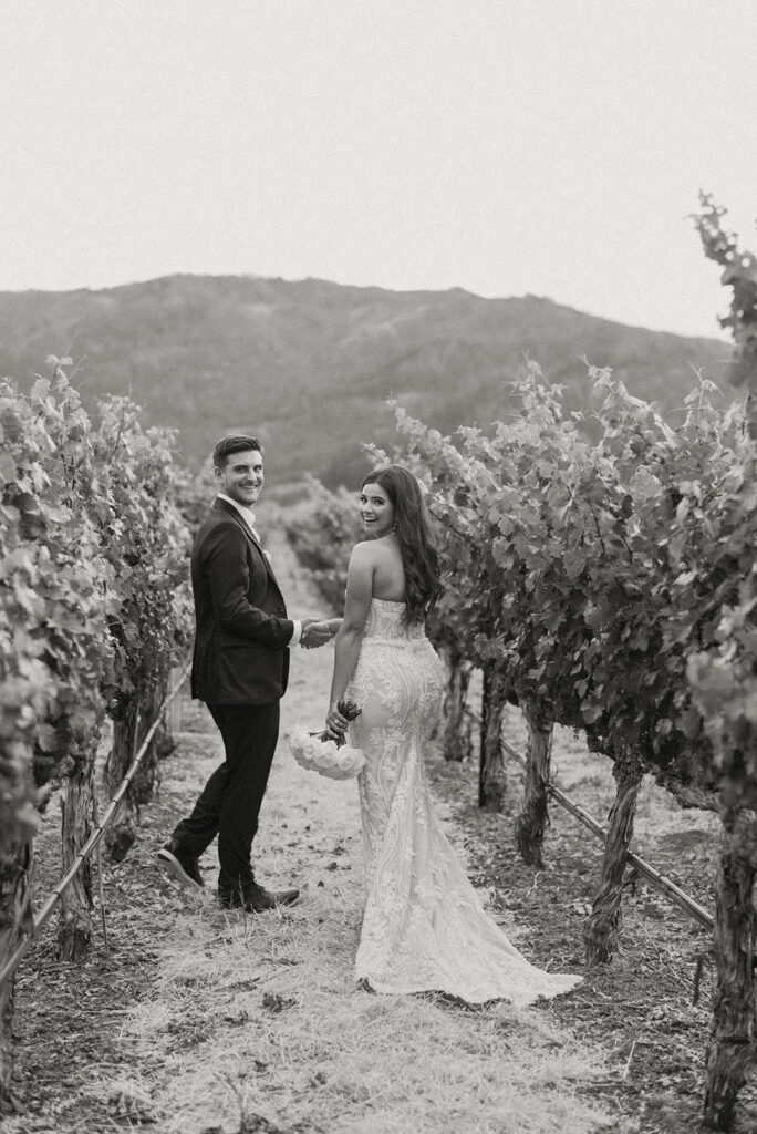 A couple dressed in wedding attire walks through a vineyard, holding hands and looking back toward the camera with mountains visible in the background.