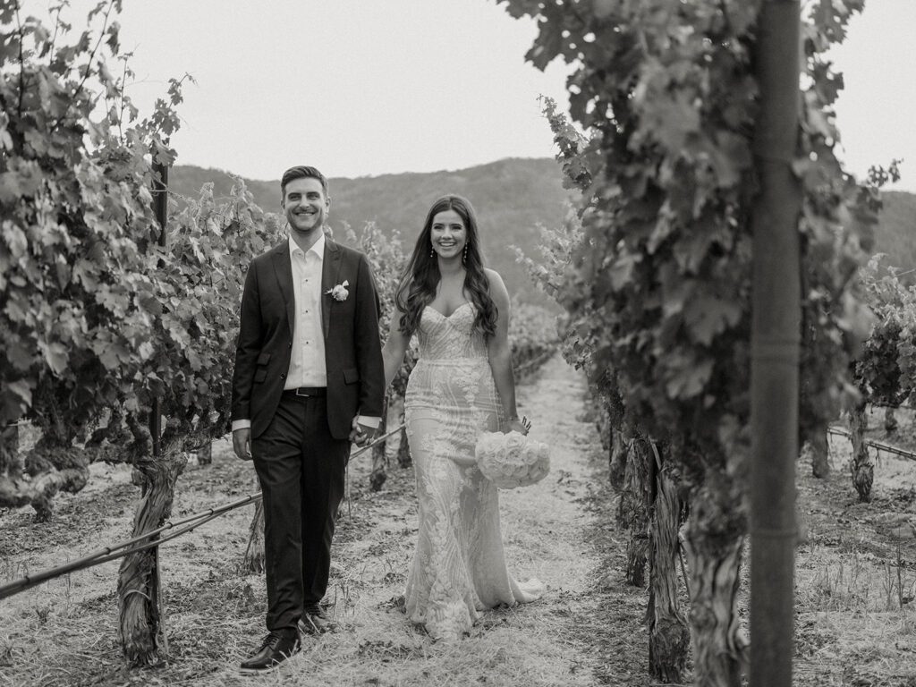 A couple dressed in wedding attire walks through a vineyard, smiling and holding hands. The background shows grapevines and distant hills.