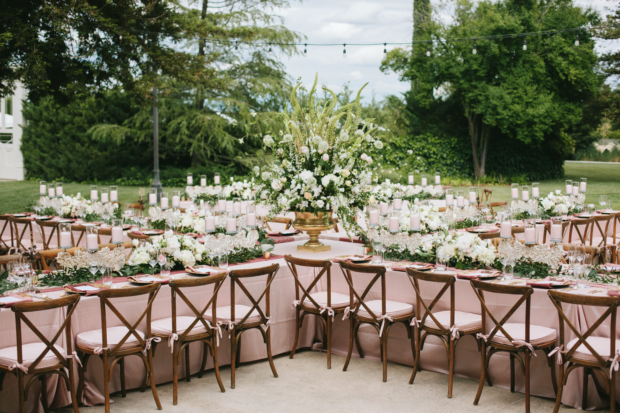 Outdoor banquet setup with wooden chairs, pink tablecloths, and white floral centerpieces surrounded by greenery. Candles decorate the tables, and string lights are overhead.