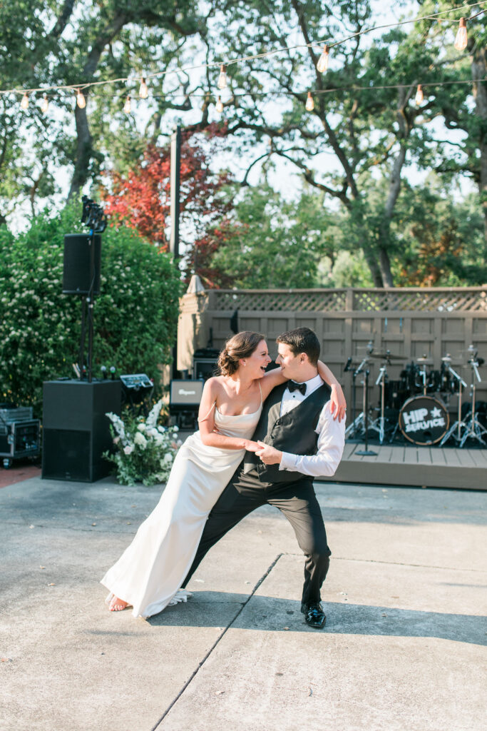 A bride and groom dance together outdoors on concrete, smiling and leaning toward each other, with a stage and musical instruments in the background.