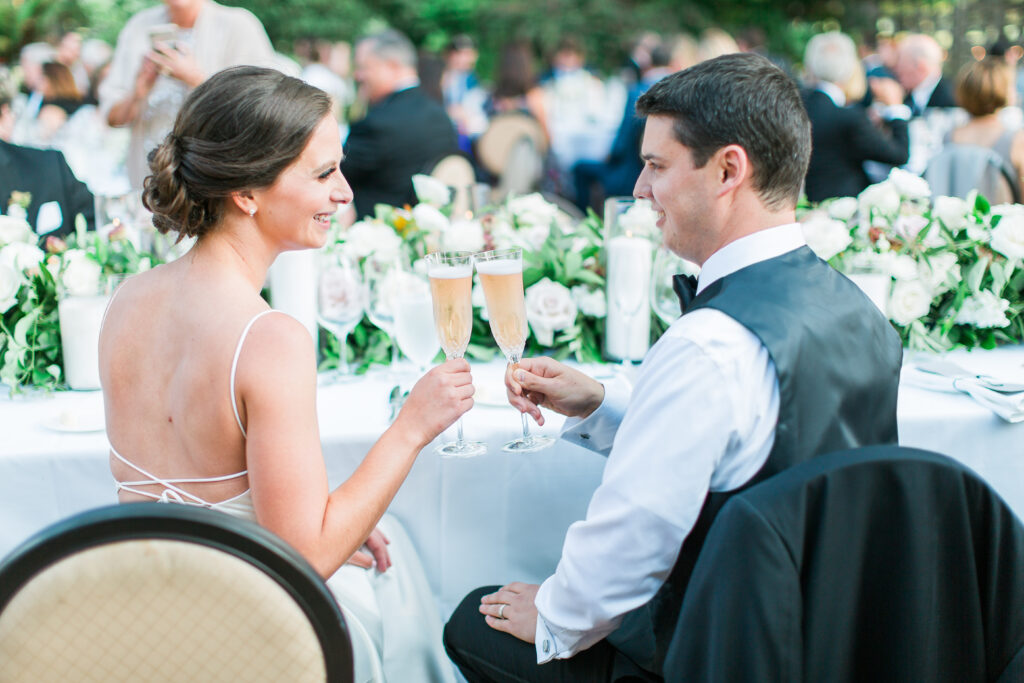 A bride and groom sit at a decorated table outdoors, smiling and clinking champagne glasses during their wedding reception.